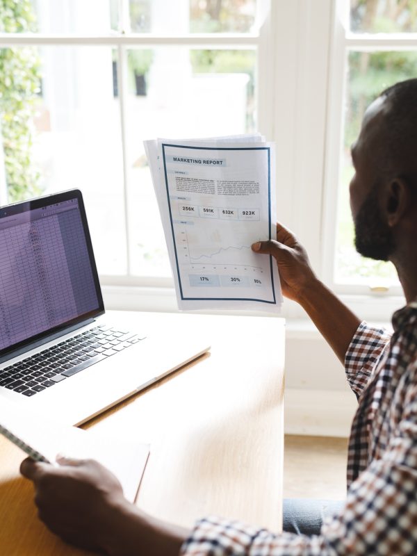 African american man sitting at table with paperwork in dining room, working remotely using laptop